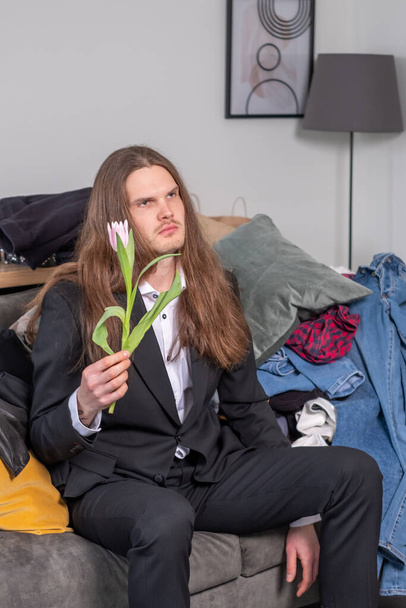 Confused, distracted and depressed man with long, brow hair looking down at something and holding a pink tuplip. Man wearing a black suit is sitting on the couch in total mess. - Фото, зображення