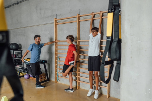 Groupe de jeunes hommes adolescents relaxant étirement et faire une pause sur les barres murales de gymnastique en bois dans la salle de gym, dans la lumière sombre, la nuit. Horizontal, vue de côté copier-espace - Photo, image
