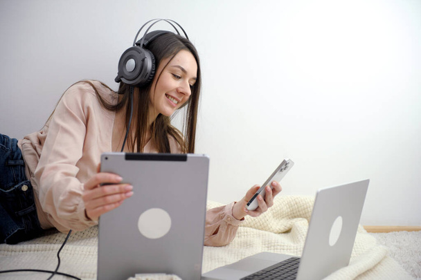 Une étudiante souriante porte un casque sans fil pour étudier en ligne avec un professeur Skype, une jeune femme heureuse apprend la langue, écoute le webinaire de la conférence, écrit des notes, regarde un ordinateur portable, enseigne à distance - Photo, image