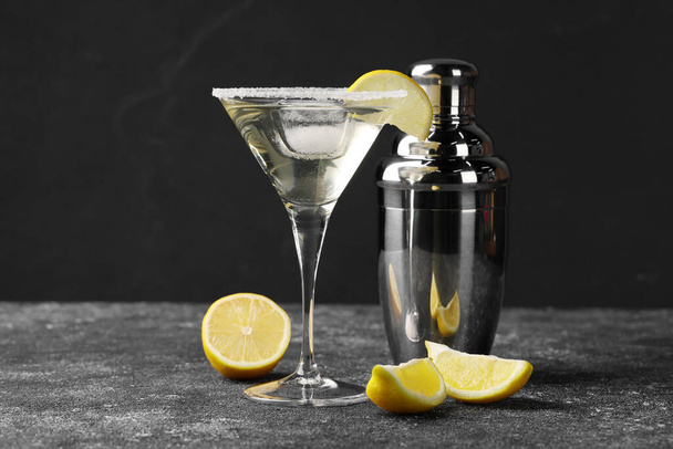 Martini glass of refreshing cocktail with lemon and ice cube near shaker on black textured table - Valokuva, kuva