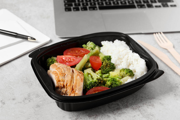 Container with tasty food, laptop, cutlery and notebook on light grey table. Business lunch - Foto, Imagem