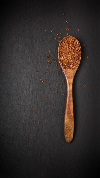 whole grain dark brown rice in a wooden spoon, also called purple rice, forbidden or chinese black rice, isolated on a black slate background, top down view with copy space - Photo, Image