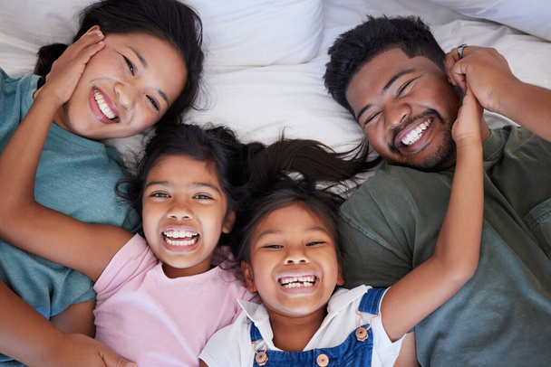 Happy, smile and portrait of family from above in bed bonding together in a bedroom at home. Happiness, love and interracial parents lying with girl children in holiday house room while on vacation - Photo, Image