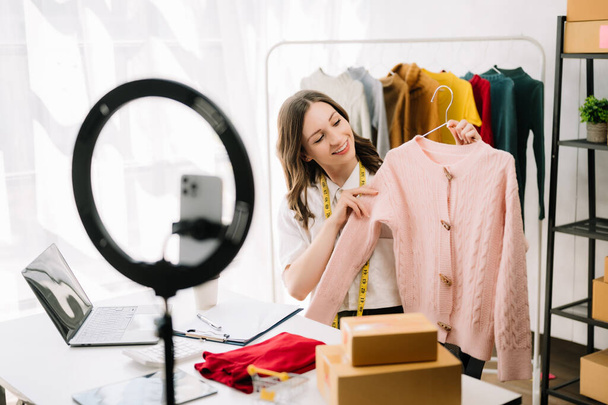 Fashion blogger concept, Young  woman selling clothes on video streaming.Startup small business SME, using smartphone or tablet taking receive and checking at office - 写真・画像