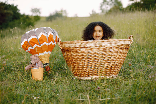 Baby girl in wicker basket with balloons in sunlight in summer time. Happy child in field on nature on a grass. African girl with black curly hair. - Foto, imagen
