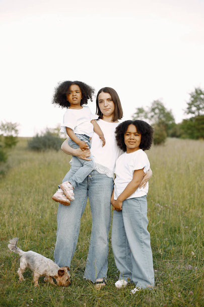 Photo of caucasian mother and two her african american daughters embracing together outdoors. Girls has black curly hair. Mother and daughers wearing white t-shirts. - Fotó, kép