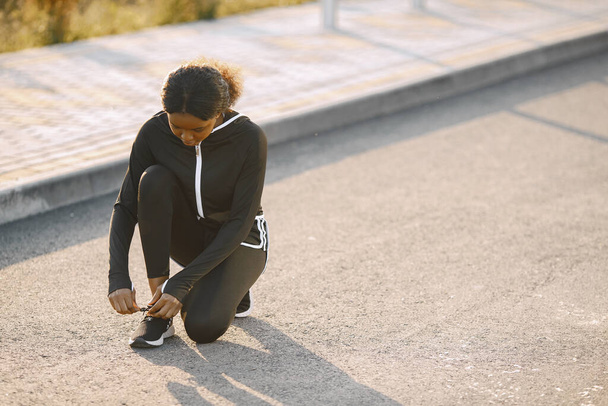 Mujer africana haciendo una pausa para atarse los cordones de los zapatos mientras trota por una carretera. Mujer usando ropa deportiva. Concepto de salud y estado físico. - Foto, imagen