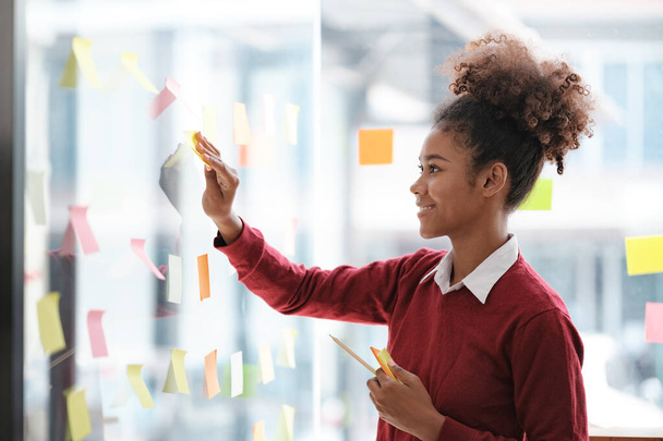 Close up of focused african American female employee write down on colorful sticky notes manage list, concentrated biracial woman work on startup brainstorm collaborate plan on stickers on glass wall - Photo, Image