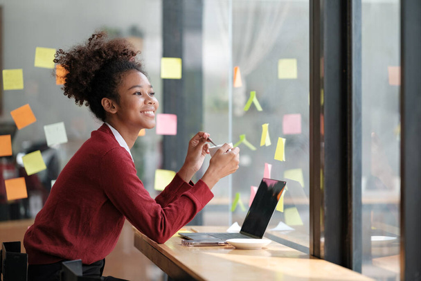 Close up of focused african American female employee write down on colorful sticky notes manage list, concentrated biracial woman work on startup brainstorm collaborate plan on stickers on glass wall - Valokuva, kuva