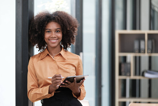 Glückliche afrikanische Geschäftsfrau, die online mit digitalen Geräten chattet, ihr Handy in der Hand, ihren Laptop benutzt, in die Kamera schaut und lächelt. Freiberufler am heimischen Arbeitsplatz mit Kopfschuss. Hohe Qualität - Foto, Bild