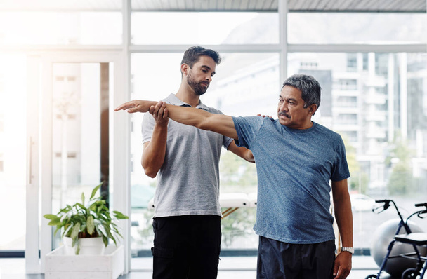 Halten Sie ihn dort, so lange Sie können. ein junger männlicher Physiotherapeut hilft einem älteren männlichen Patienten mit Bewegungsübungen in einer Klinik - Foto, Bild