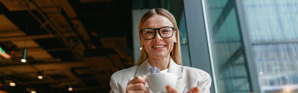 Bizneswoman w okularach po przerwie na kawę w ciągu dnia pracy w kawiarni i patrząc w kamerę - Zdjęcie, obraz