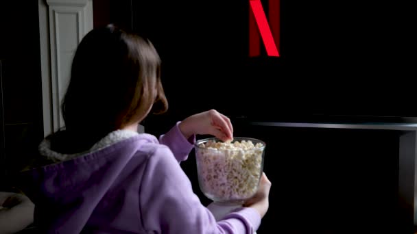 tiener meisje zit op de vloer en kijkt netflix op groot scherm van TV screensaver slow motion flitsen op zwarte achtergrond in rood blauw in handen van jonge vrouw popcorn meisje neemt het en eet het betoverd - Video
