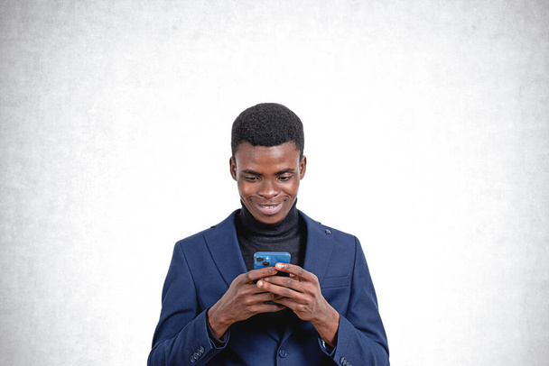 Portrait of cheerful young African American businessman using smartphone standing over concrete wall background. Concept of communication - Photo, image