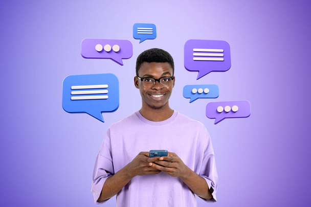 Веселий молодий афроамериканець у окулярах, користуючись смартфоном над фіолетовим фоном з бульбашками мови. Концепція соціальних медіа і балачок. - Фото, зображення
