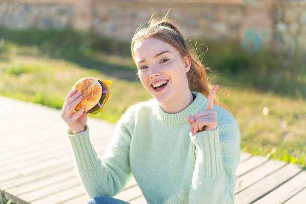 Genç, güzel bir kız elinde hamburgerle harika bir fikri işaret ediyor. - Fotoğraf, Görsel