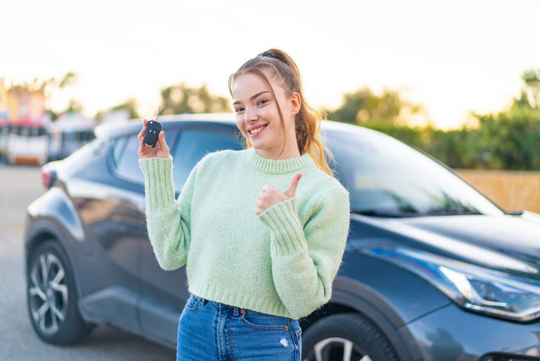 Nuori kaunis tyttö pitelee auton avaimet ulkona peukalot pystyssä, koska jotain hyvää on tapahtunut - Valokuva, kuva