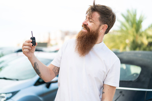 Redhead man with beard holding car keys at outdoors with happy expression - Photo, image