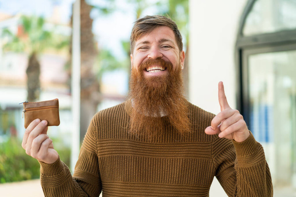Rothaariger Mann mit Bart hält Geldbörse im Freien und zeigt auf eine großartige Idee - Foto, Bild