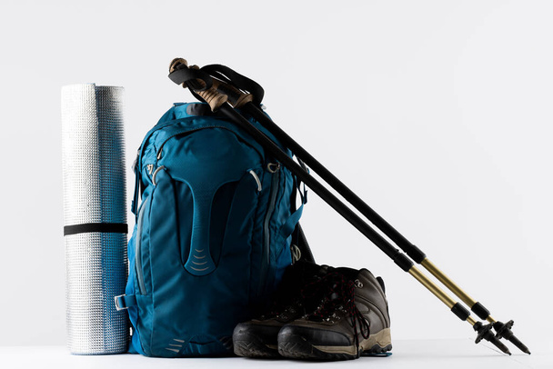 Close up of blue backpack, mat, trekking shoes and sticks on white background with copy space. National camping month, equipment and celebration concept. - Foto, Imagen