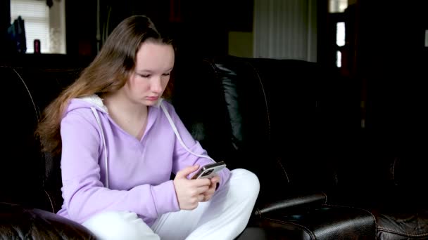 Hymyilevä teini-ikäinen tyttö rentoutuu, istuu sohvalla kotona matkapuhelimella, keskustele ystävän kanssa, onnellinen nuori nainen sohvalla, pidä älypuhelin verkkokaupassa tai tarkista mobiilisovellus tai sosiaaliset verkostot. - Materiaali, video