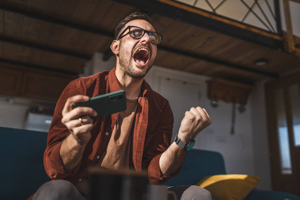 Un homme caucasien adulte assis à la maison sourire heureux jouer à des jeux vidéo loisirs s'amuser tenir téléphone mobile smartphone avoir plaisir à copier espace porter des lunettes et chemise - Photo, image