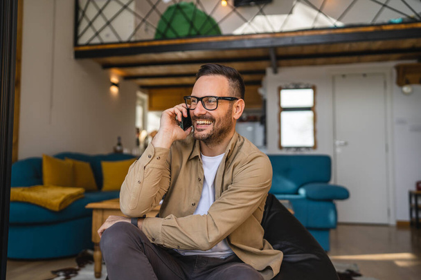 Um homem adulto caucasiano masculino usar smartphone telefone celular para fazer uma chamada falar sorriso feliz enquanto se senta no sofá-cama em casa usar camisa e óculos espaço cópia pessoa real - Foto, Imagem