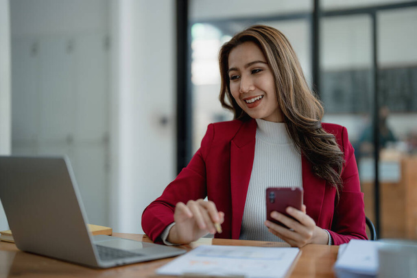 Retrato de una joven asiática que muestra una cara sonriente mientras usa su teléfono, computadora y documentos financieros en su escritorio en las primeras horas de la mañana. - Foto, imagen