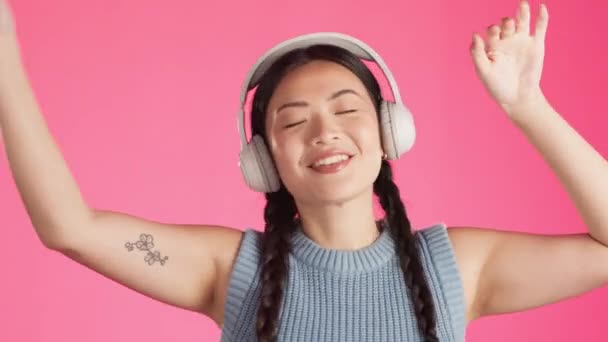 Happy woman, dancing and music headphones on isolated studio background for energy, freedom or expression. Smile, asian and fashion dancer listening to radio, podcast or audio on pink party backdrop. - Footage, Video