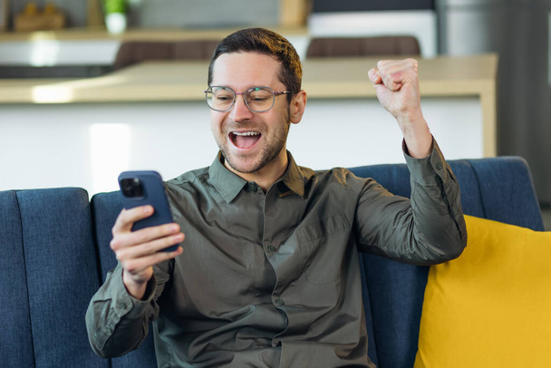 Young handsome man using smartphone sitting on the sofa at home very happy and excited doing winner gesture with arms raised, smiling and screaming for success. celebration concept. - Foto, Imagen