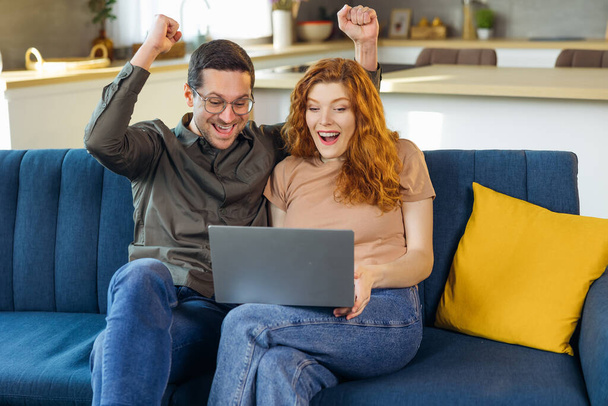 Şok olmuş genç aile çifti koltukta sarılıp el kaldırıyor çığlık çığlığa e-postaları okuyor ve dizüstü bilgisayardan harika haberler geliyor. Şanslı karı piyango kazanmasını kutluyor. Satın almak için harika bir teklif alıyor. - Fotoğraf, Görsel