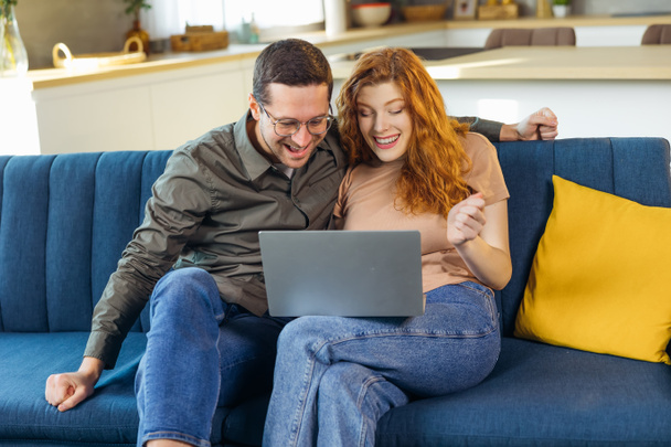 Iloinen pari istua rentoutua kodikas sohvalla tarkastella kannettavan tietokoneen näytön voittoarpajaiset verkossa, onnellinen biracial aviomies ja vaimo tuntuu innoissaan euforinen hyviä uutisia tietokoneella - Valokuva, kuva