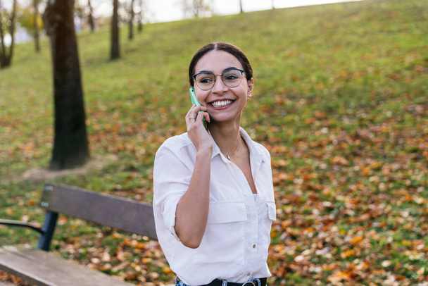 Een jonge blanke vrouw in een casual outfit en bril staat in een park, verdiept in een telefoongesprek als de zon achter haar ondergaat. Het warme licht baadt de scène in een gouden gloed. - Foto, afbeelding