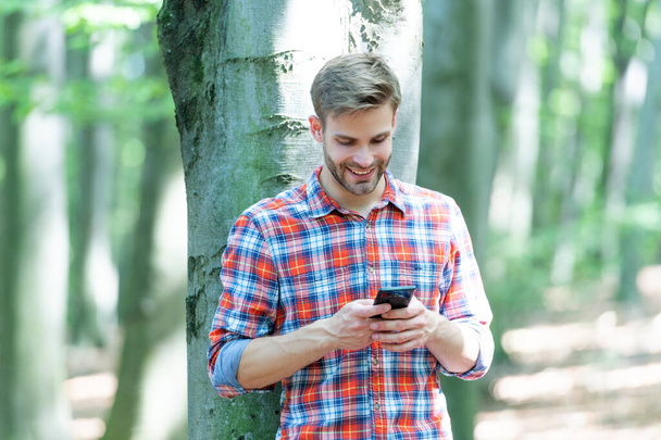cheerful man messaging on phone outdoor. man messaging on phone outside. man messaging on phone wearing checkered shirt. photo of man messaging on phone in the forest. - Φωτογραφία, εικόνα