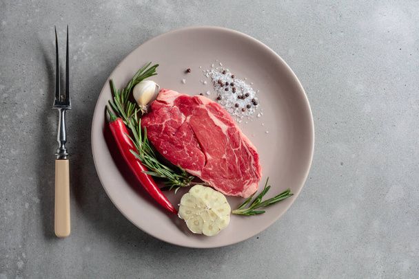 Raw beef steak for grilling with rosemary, garlic, salt, and pepper. Grey concrete background. Top view. - Foto, imagen