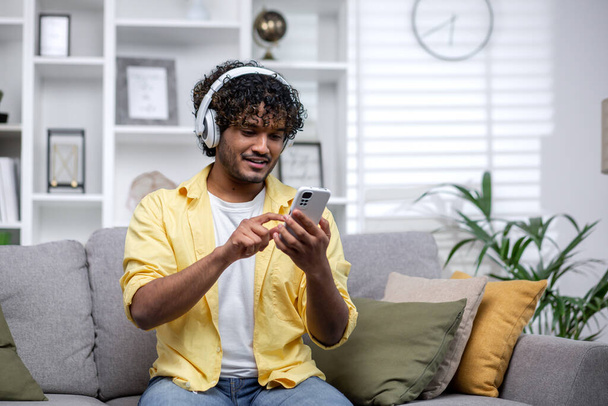 Junge lächelnde hispanische Mann zu Hause Musik hören mit Online-App auf dem Handy, Mann lächelt allein zu Hause auf der Couch im Wohnzimmer sitzen Streaming-Video. - Foto, Bild