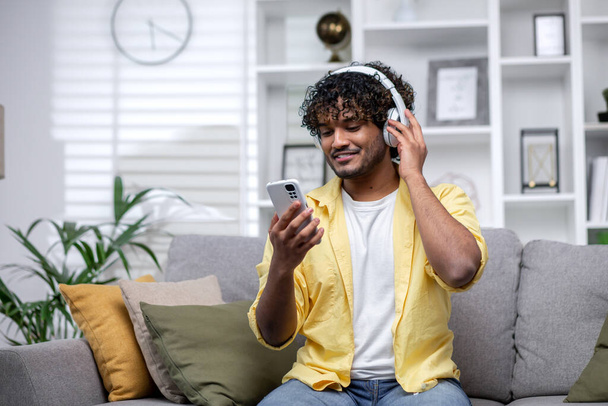 Young smiling hispanic man at home listening to music using online app on phone, man smiling alone at home sitting on couch in living room watching streaming video. - Fotoğraf, Görsel