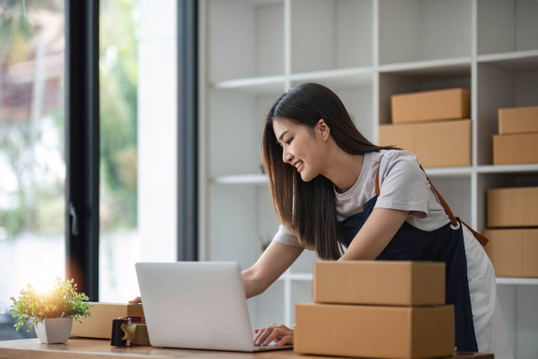 A portrait of a young Asian woman, e-commerce employee sitting in the office full of packages in the background write note of orders and a calculator, for SME business ecommerce and delivery business - Photo, Image