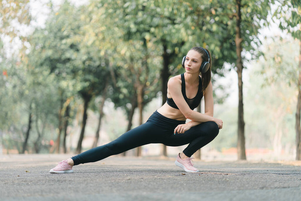 Een jonge vrouwelijke atleet, gekleed voor hardlopen, wordt getoond strekken haar benen ter voorbereiding op een jog door het park - Foto, afbeelding