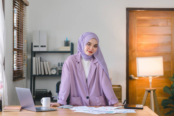 Portret pięknej muzułmańskiej milenijnej azjatyckiej bizneswoman w fioletowym hidżabie pracującej zdalnie z jej biura. Patrzę w kamerę.. - Zdjęcie, obraz