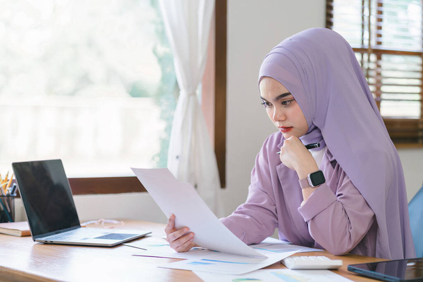 A young Muslim businesswoman wearing a purple hijab is working from home and appears stressed while using her laptop, looking worn out and anxious - Valokuva, kuva