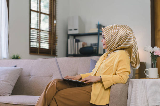 Портрет прекрасной азиатской бизнесвумен тысячелетия, носящей хиджаб, работающей удаленно от своего домашнего офиса, сидя на диване - Фото, изображение