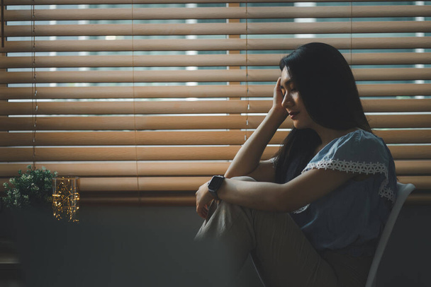 Traurige und deprimierte junge Frau, die vor dem Fenster im Wohnzimmer sitzt, mit traurigem Gesichtsausdruck nach draußen blickt und Gefühle der Erschöpfung, Einsamkeit und des Unglücks vermittelt - Foto, Bild