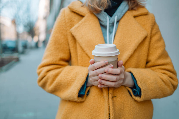 Charming happy young woman dressed in a bright yellow coat holds coffee to go. Girl spend free time traveling in european old town enjoying cup of tea, coffee.  - Foto, afbeelding