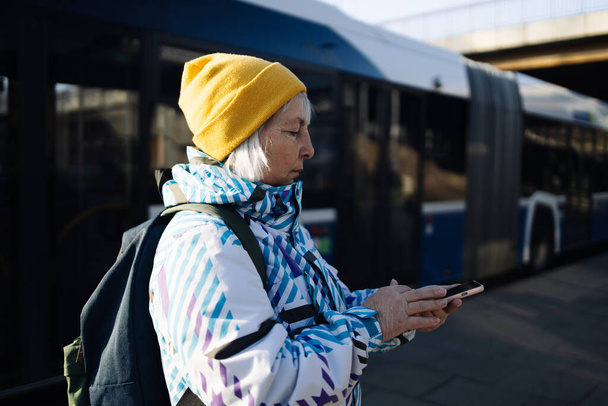Vanha 60-luvun nainen matkatavaroineen seuraa junan aikataulua ja ostaa junalippuja älypuhelimellaan sähköisellä sovelluksella odottaessaan asemalla. - Valokuva, kuva