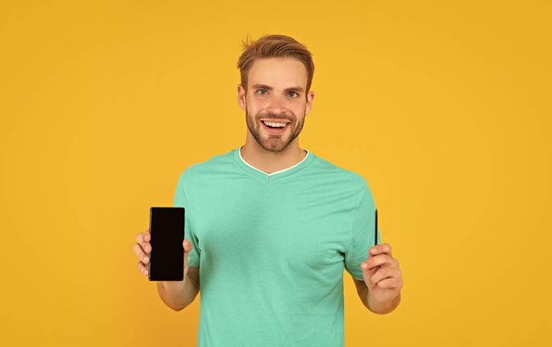 happy guy showing smartphone on yellow background. product proposal. advertisement presentation of digital pen. copy space. man presenting phone screen and stylus. - Fotografie, Obrázek