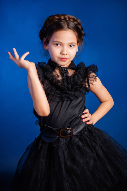 Portrait of a little girl in a black dress with a pigtail hairstyle on her head poses, isolated on a dark background with blue backlight.  - Photo, Image