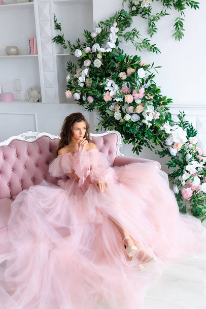 Young brunette girl with long beautiful hair in a lush pink tulle dress in a light white studio on the background of shelves with books, a fireplace and flower arrangements. High quality photo - Foto, immagini