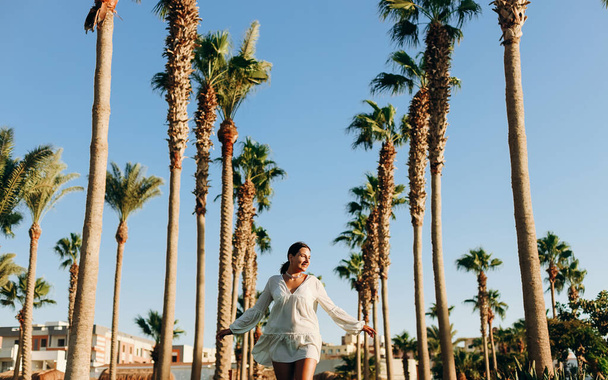 A beautiful young happy girl enjoys a sunny day against the background of palm trees and blue sky. Horizontal photo - Photo, Image