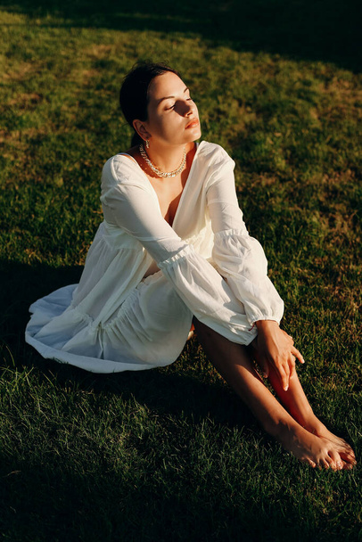 A beautiful young happy girl enjoys a sunny day on the green grass, in a white dress. Vertical photo - Фото, зображення